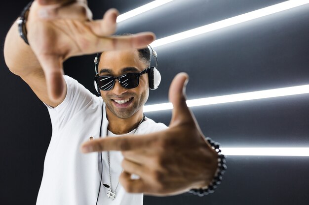 Joven guapo sonriente hipster negro en traje blanco escuchando música en auriculares y bailando al estilo hip hop en discoteca, divirtiéndose