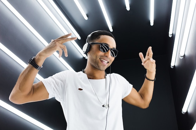 Joven guapo sonriente hipster negro en traje blanco escuchando música en auriculares y bailando al estilo hip hop en discoteca, divirtiéndose