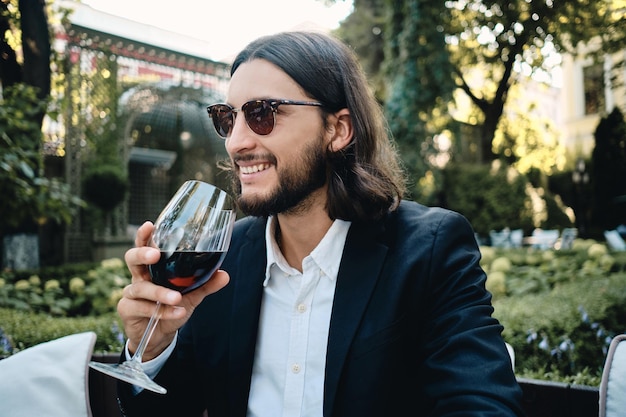 Joven guapo sonriente moreno barbudo felizmente bebiendo vino tinto en el restaurante al aire libre