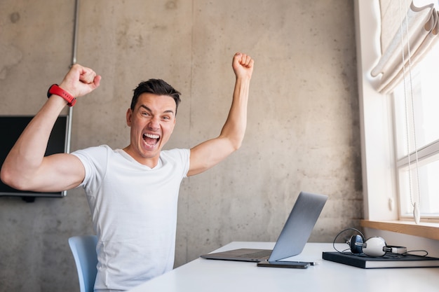 Foto gratuita joven guapo sonriente en traje casual sentado en la mesa trabajando en la computadora portátil, autónomo en casa, levantando las manos en el éxito