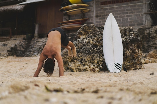 Un joven y guapo surfista en la orilla del océano está calentando antes de surfear. ejercicios antes del deporte, estiramientos antes del surf.