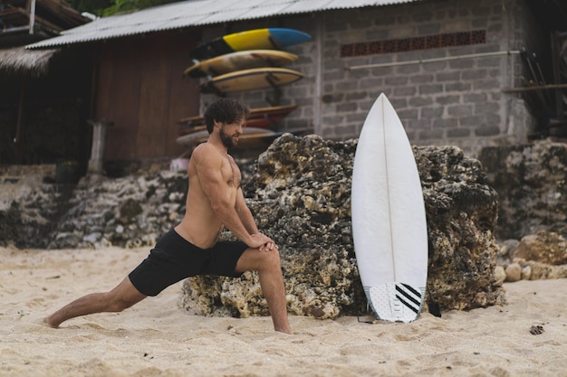 Un joven y guapo surfista en la orilla del océano está calentando antes de surfear. ejercicios antes del deporte, estiramientos antes del surf.