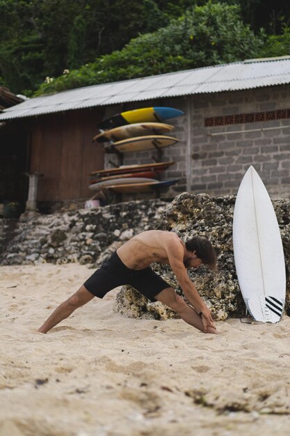 Un joven y guapo surfista en la orilla del océano está calentando antes de surfear. ejercicios antes del deporte, estiramientos antes del surf.