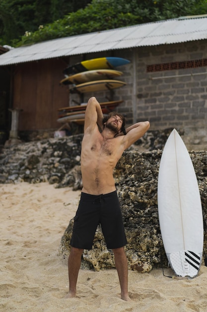 Un joven y guapo surfista en la orilla del océano está calentando antes de surfear. ejercicios antes del deporte, estiramientos antes del surf.