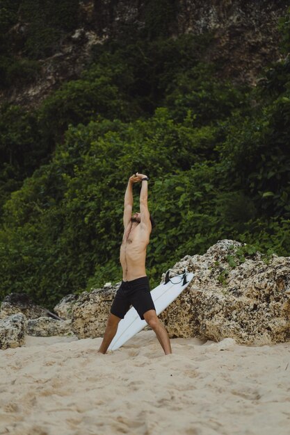 Un joven y guapo surfista en la orilla del océano está calentando antes de surfear. ejercicios antes del deporte, estiramientos antes del surf.