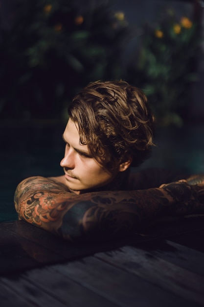 joven guapo en tatuajes descansando en la piscina al aire libre.