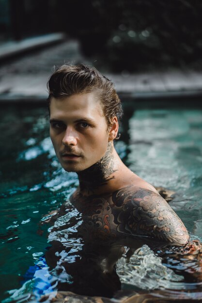 joven guapo en tatuajes descansando en la piscina al aire libre.