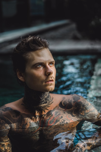 joven guapo en tatuajes descansando en la piscina al aire libre.