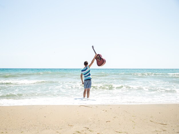 Joven con guitarra acústica en la playa, emociones alegres, el concepto de ocio y música
