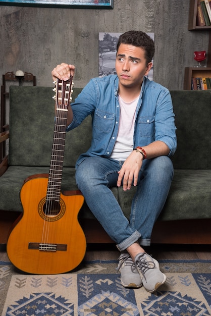 Joven guitarrista sosteniendo una hermosa guitarra y sentado en el sofá. Foto de alta calidad