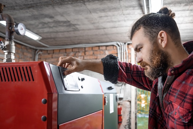 Un joven en una habitación con una caldera de combustible sólido, trabajando en biocombustible, calefacción económica.