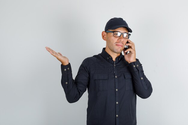 Joven hablando por teléfono móvil en camisa negra con gorra, gafas