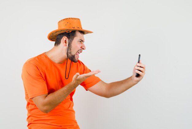 Joven hablando por video chat en camiseta naranja, sombrero y mirando enojado, vista frontal.