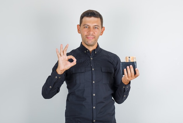 Joven haciendo bien firmar con billetera en camisa negra y mirando feliz