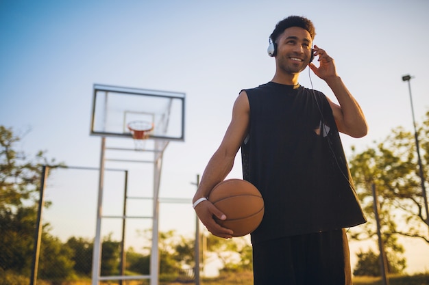 Joven haciendo deporte, jugando baloncesto al amanecer, escuchando música en auriculares