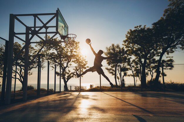 Joven haciendo deporte, jugando baloncesto al amanecer, saltando silueta