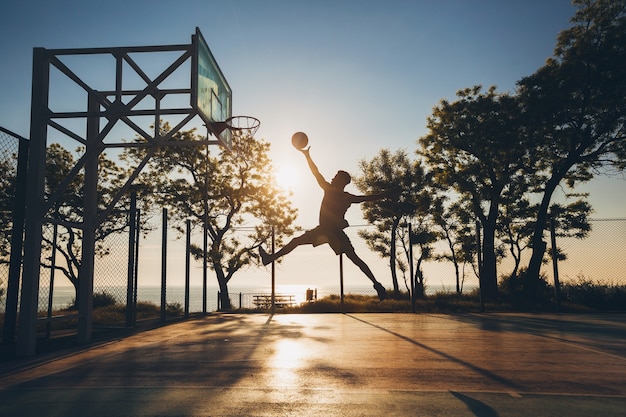 Joven haciendo deporte, jugando baloncesto al amanecer, saltando silueta