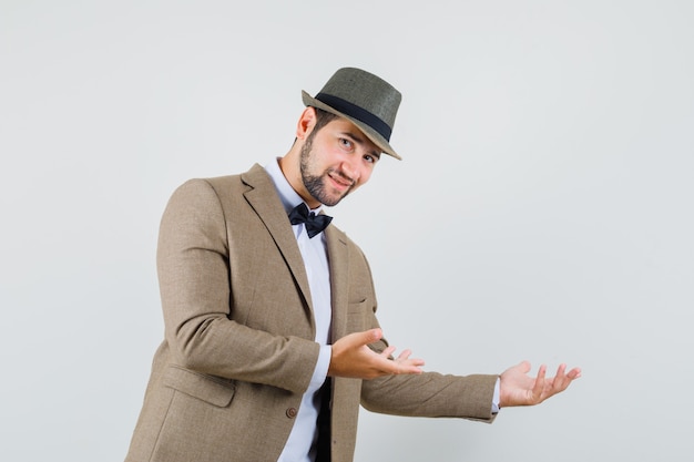 Foto gratuita joven haciendo gesto de bienvenida cortésmente en traje, sombrero y mirando suave, vista frontal.