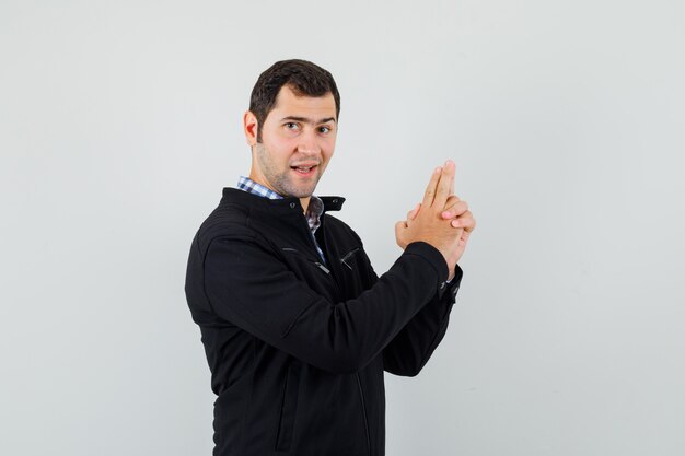 Joven haciendo pistola de dedo firmar en camisa, chaqueta y mirando confiado