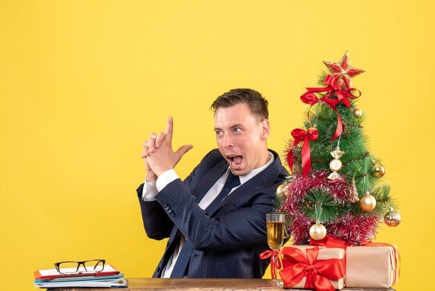 Joven haciendo pistola de dedo sentado en la mesa cerca del árbol de Navidad y presenta en amarillo