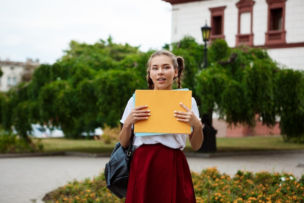 Joven hermosa alegre estudiante sonriente, sosteniendo carpetas al aire libre