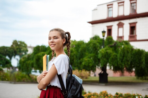 Joven hermosa alegre estudiante sonriente, sosteniendo carpetas al aire libre