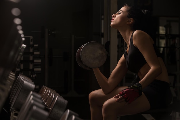 Joven hermosa atleta rubia sexy ejercicios de culturista en el gimnasio en hermosa ropa deportiva sobre un fondo oscuro