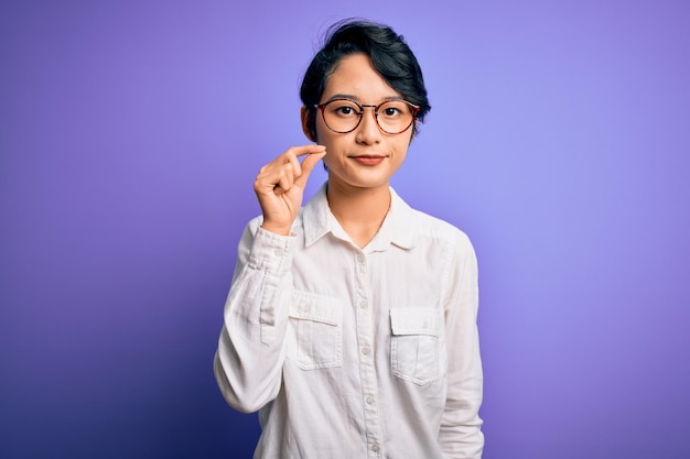 Foto gratuita joven hermosa chica asiática con camisa casual y anteojos de pie sobre un fondo morado boca y labios cerrados como zip con los dedos tabú secreto y silencioso hablando