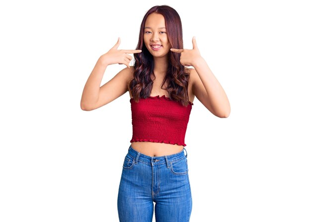 Joven hermosa chica china con ropa informal sonriendo alegre mostrando y señalando con los dedos los dientes y la boca. concepto de salud dental.