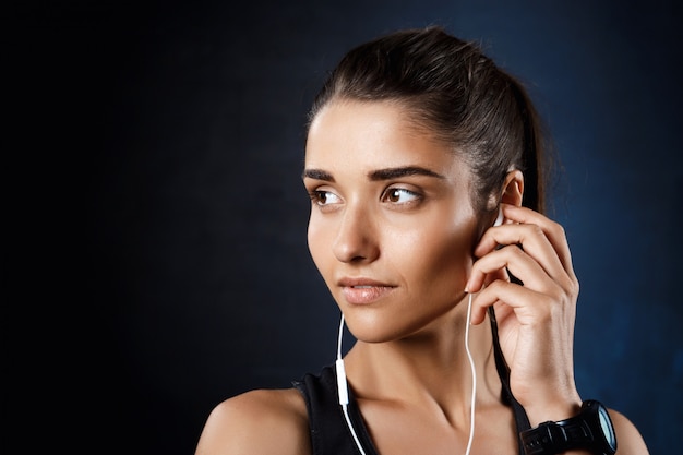 Joven hermosa chica deportiva escuchando música sobre la pared oscura.