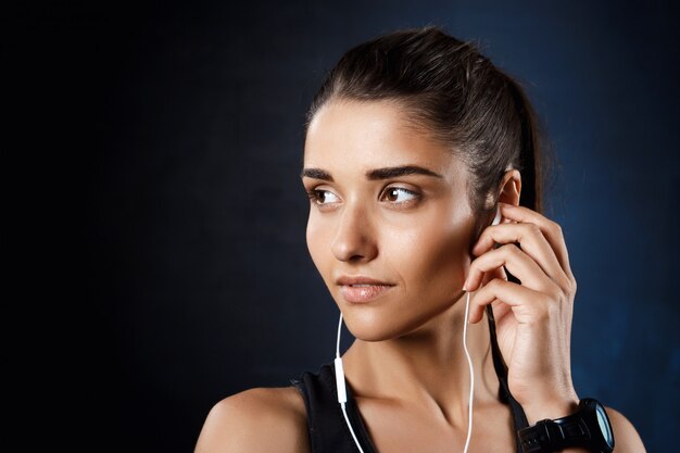 Joven hermosa chica deportiva escuchando música sobre la pared oscura.