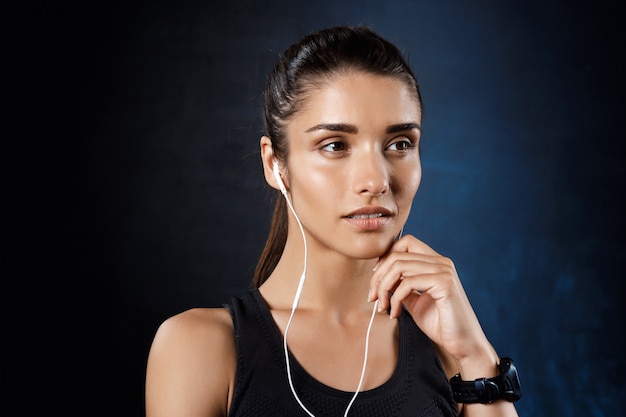 Joven hermosa chica deportiva escuchando música sobre la pared oscura.