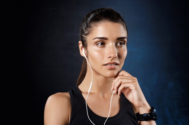 Joven hermosa chica deportiva escuchando música sobre la pared oscura.