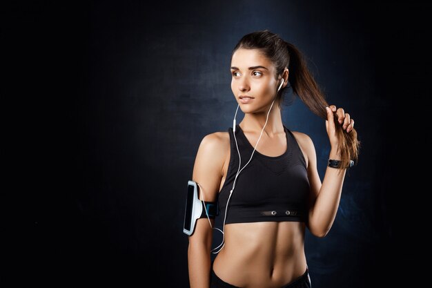 Joven hermosa chica deportiva escuchando música sobre la pared oscura.