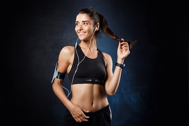 Joven hermosa chica deportiva escuchando música sobre la pared oscura.