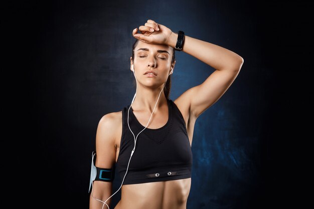 Joven hermosa chica deportiva escuchando música sobre la pared oscura.