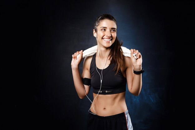 Joven hermosa chica deportiva escuchando música sobre la pared oscura.