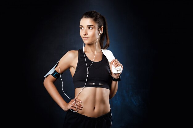 Joven hermosa chica deportiva escuchando música sobre la pared oscura.