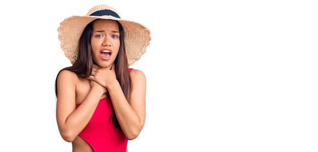 Joven hermosa chica latina con traje de baño y sombrero de verano gritando y asfixiándose por el doloroso estrangulamiento. problema de salud. concepto de asfixia y suicidio.