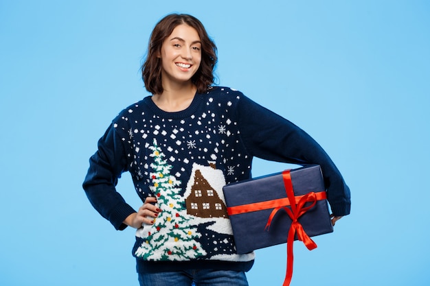 Joven hermosa chica morena en suéter de punto acogedor sonriendo sosteniendo la caja de regalo sobre la pared azul