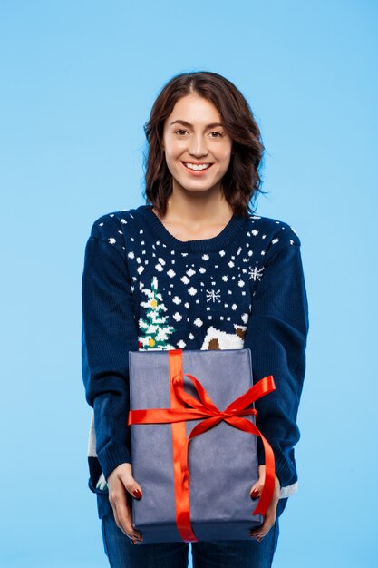 Joven hermosa chica morena en suéter de punto acogedor sonriendo sosteniendo la caja de regalo sobre la pared azul