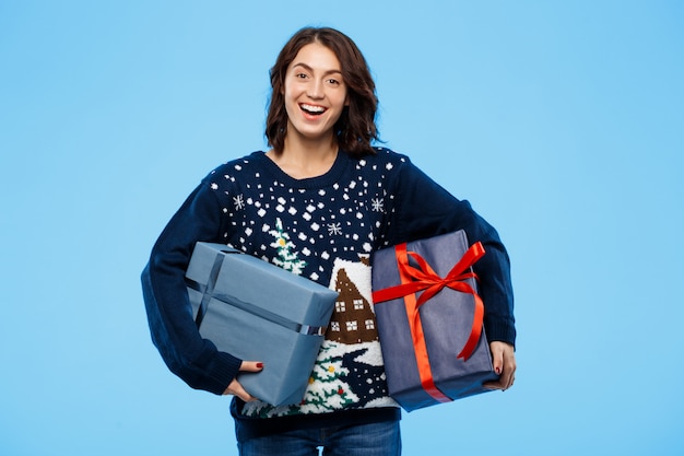 Joven hermosa chica morena en suéter de punto acogedor sonriendo sosteniendo cajas de regalo sobre pared azul