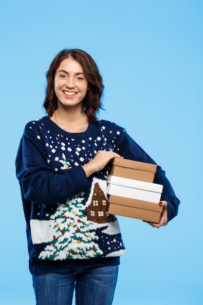 Joven hermosa chica morena en suéter de punto acogedor sonriendo sosteniendo cajas de regalo sobre pared azul