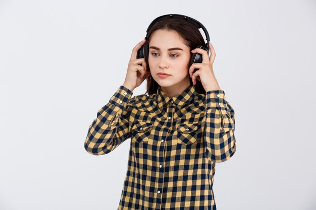 Joven hermosa chica morena vestida con camisa a cuadros escuchando música en auriculares mirando a otro lado sobre la pared blanca