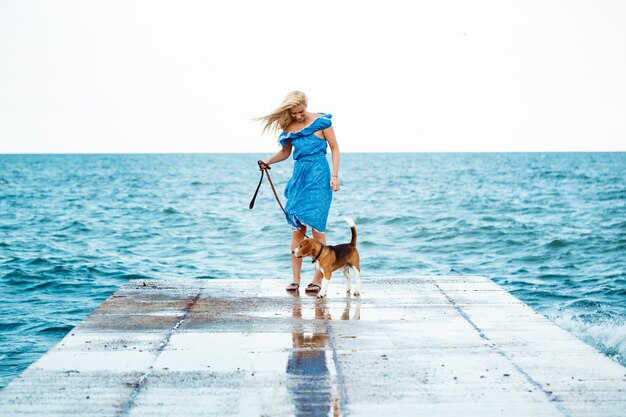 Joven hermosa chica rubia caminando, jugando con el perro beagle en la playa.