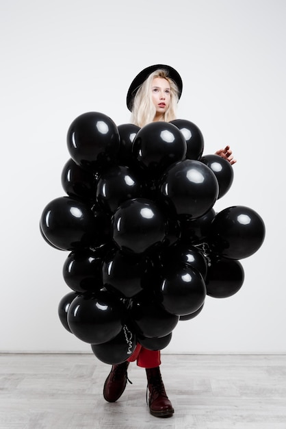 Joven hermosa chica rubia sosteniendo globos negros sobre pared blanca