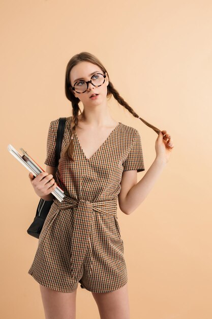 Joven hermosa chica soñadora con dos trenzas en traje de tweed y anteojos con mochila negra en el hombro sosteniendo un libro y libretas en la mano mientras mira cuidadosamente hacia arriba sobre un fondo beige