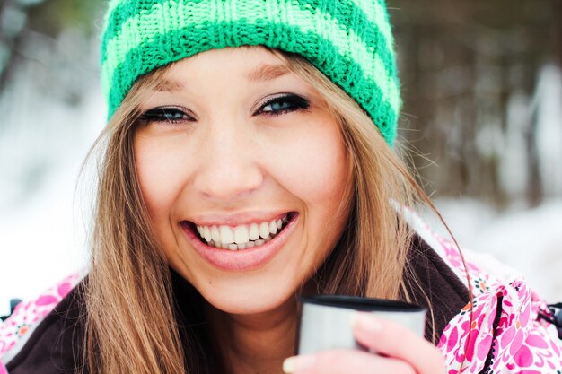 Joven hermosa chica sonriente en una chaqueta carmesí y sombrero verde beber té caliente de un termo en las montañas cubiertas de nieve