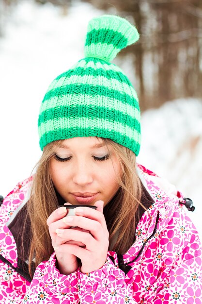 Joven hermosa chica sonriente en una chaqueta carmesí y sombrero verde beber té caliente de un termo en las montañas cubiertas de nieve