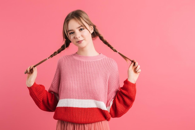 Foto gratuita joven hermosa dama en suéter sosteniendo dos trenzas de pelo en las manos mientras mira felizmente en cámara sobre fondo rosa